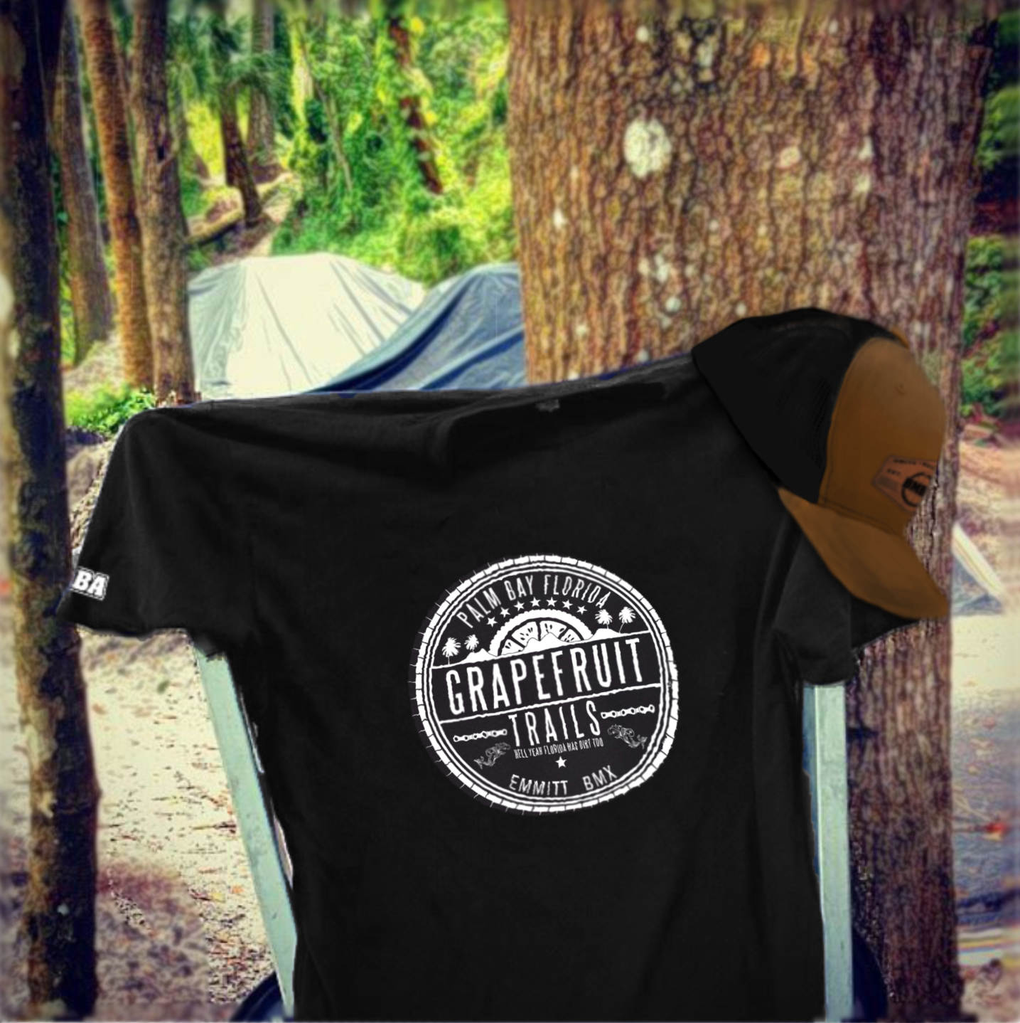Black t-shirt with 'Grapefruit Trails' logo and trucker hat on a wheelbarrow in a forest setting with dirt jumps in background.